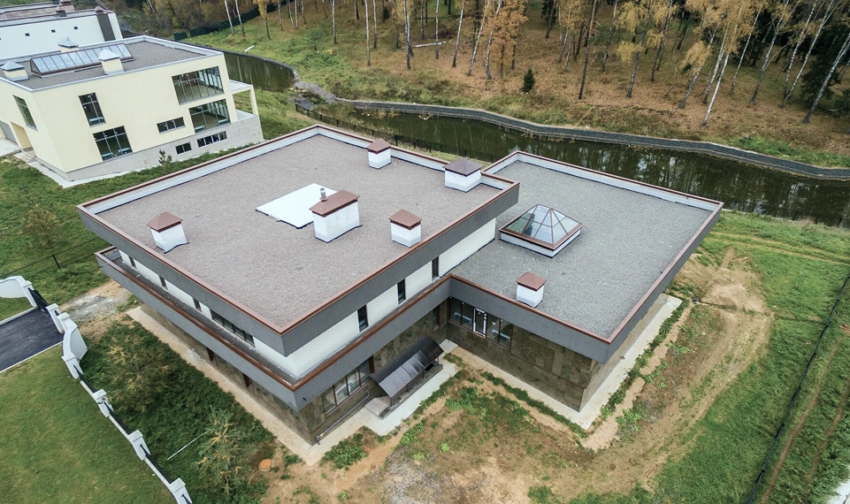 Aerial view of a modern two-story building with a flat roof, surrounded by greenery and a calm waterway, showcasing contemporary architecture.